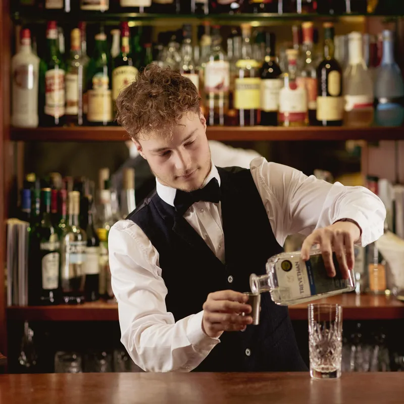 Bartender in a waistcoat pouring gin into a jigger behind a wooden bar