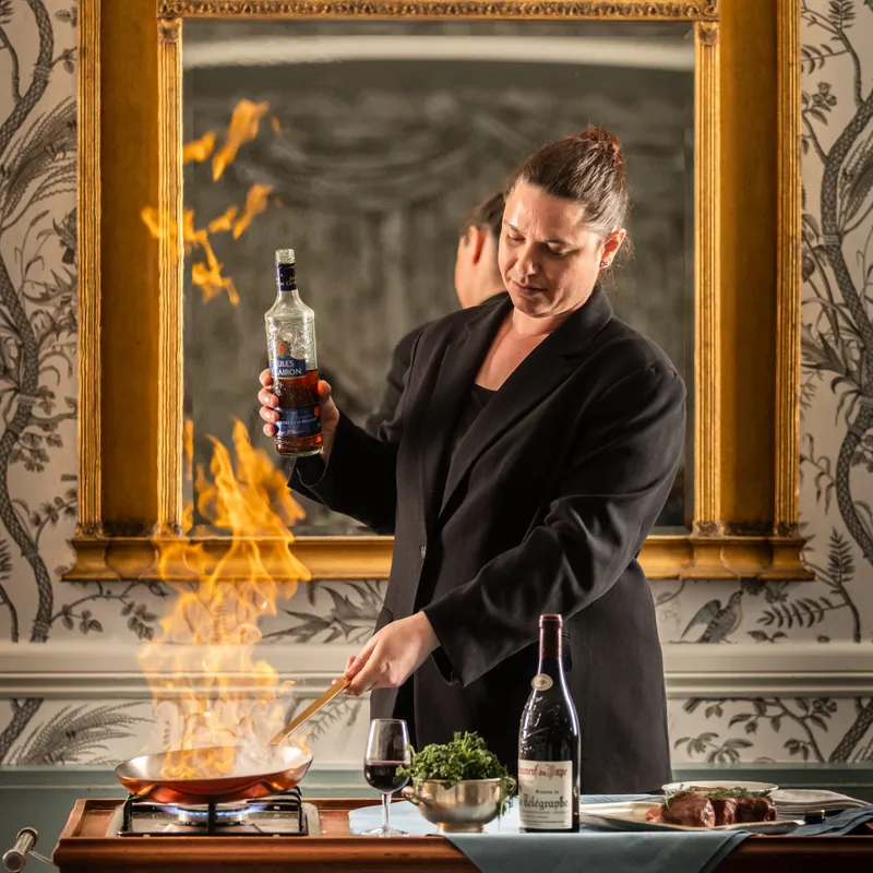 Chef flambéing food in a frying pan while holding a brandy bottle beside a table set with wine and steak.