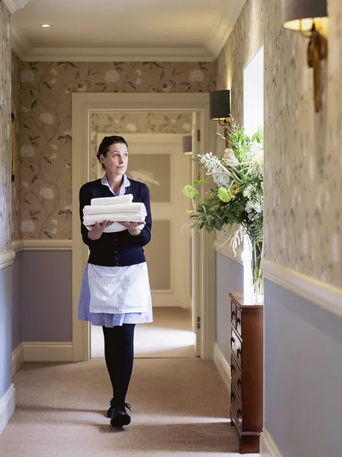 Housekeeper carrying a stack of folded white towels through a softly lit, floral-wallpapered hallway.