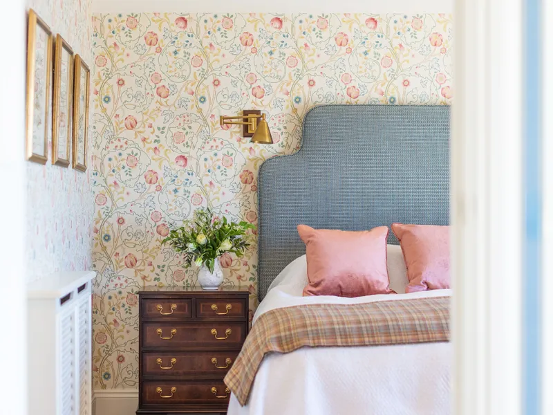 Elegant bedroom with floral wallpaper, a blue upholstered headboard, pink cushions and a dark wooden bedside chest.