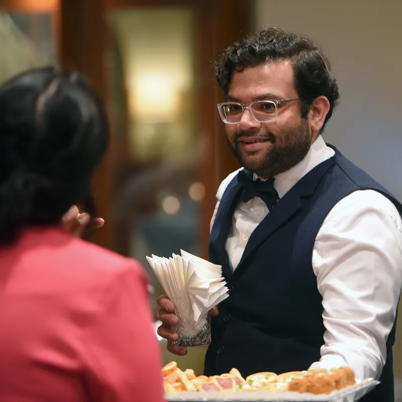 Man in a navy waistcoat and bow tie holds folded napkins while speaking with a guest at a reception.