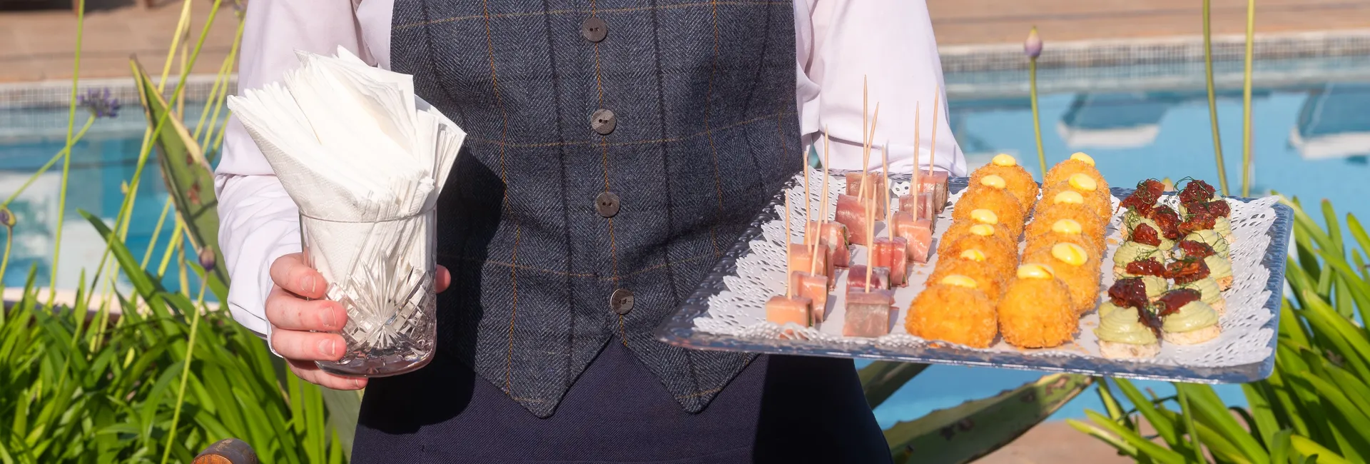 A smiling server holds a tray of canapés and a glass of napkins beside an outdoor swimming pool.