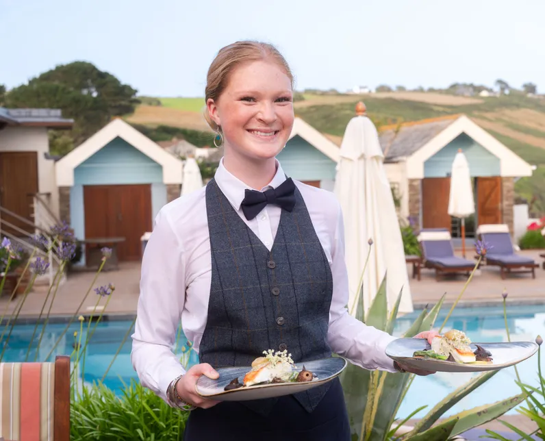 Smiling waitress holding two plated dishes beside an outdoor swimming pool