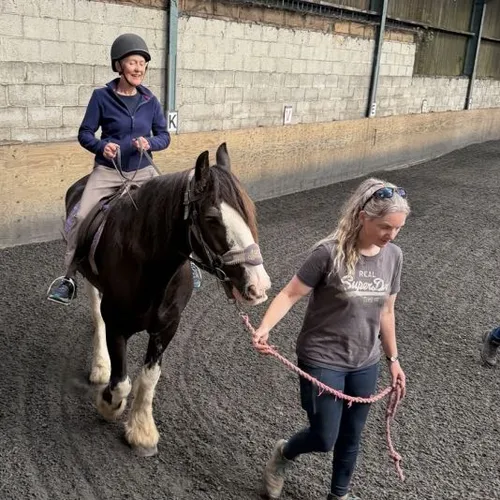A woman in a riding helmet sits on a black and white horse while another woman leads it through an indoor riding arena.