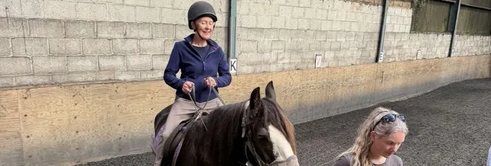 A woman in a riding helmet sits on a black and white horse while another woman leads it through an indoor riding arena.