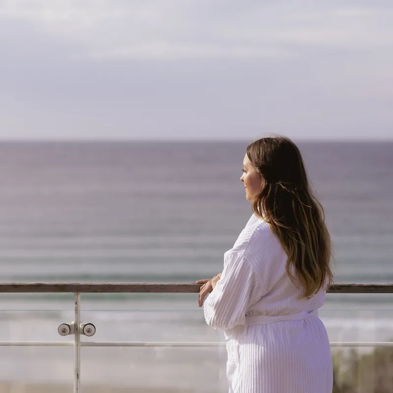 Woman in a white dressing gown leaning on a balcony railing and looking out to sea