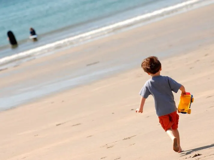 Child running barefoot across a sandy beach towards the sea, carrying a yellow toy truck
