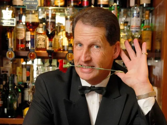 Man in a black tuxedo holding a red flower between his teeth and raising his hand in front of a bar