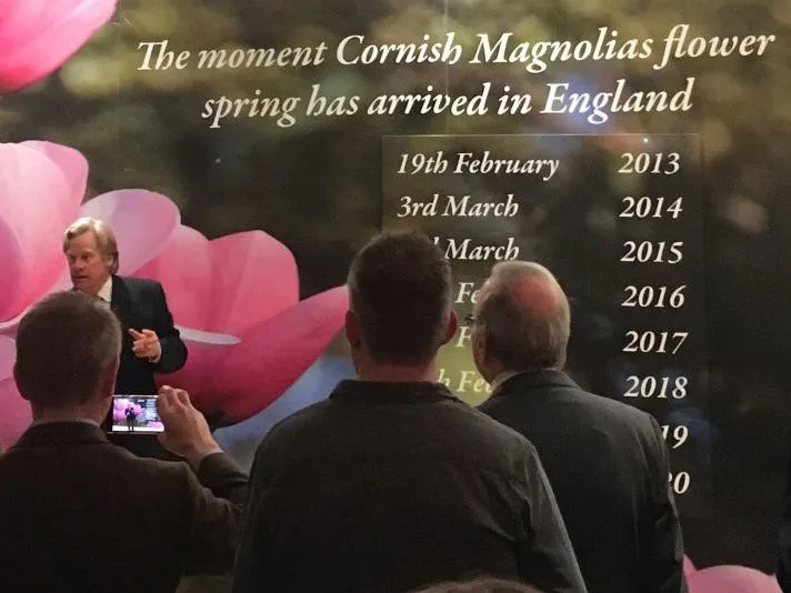 A speaker addresses an audience beside a display about Cornish magnolias flowering and the arrival of spring in England.