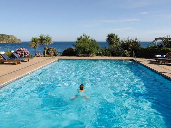 Person swimming in a sunlit outdoor pool overlooking the sea, with loungers, palm trees and flowering shrubs nearby.