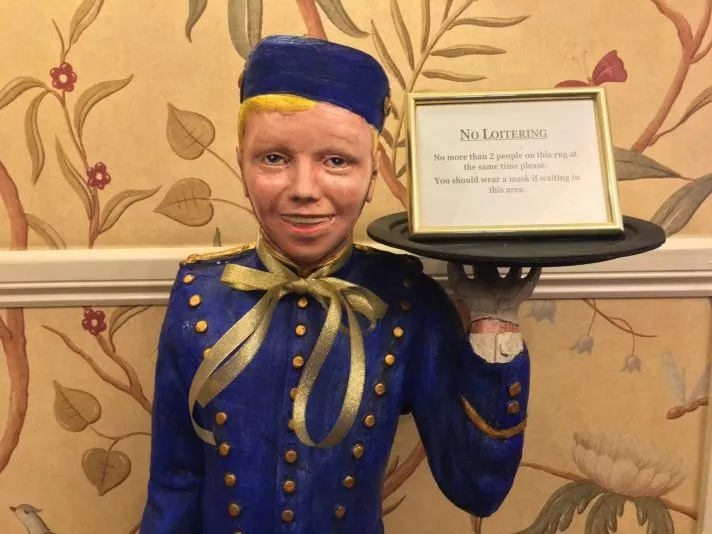 Bellhop mannequin in a blue uniform holding a framed no-loitering notice on a tray