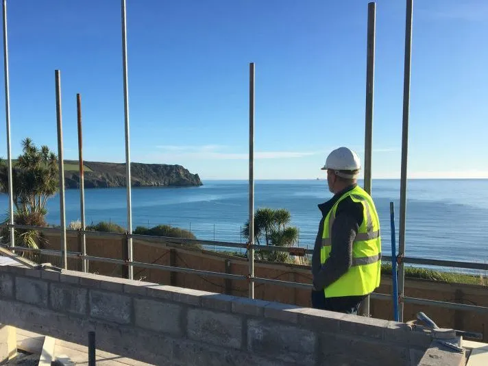 Construction worker in a white hard hat and high-visibility vest overlooking the sea from a coastal building site