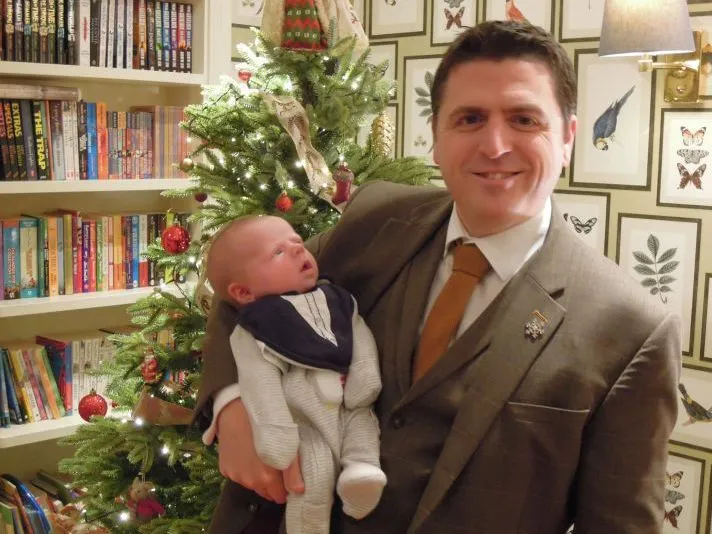 Smiling man in a brown suit holding a baby beside a decorated Christmas tree