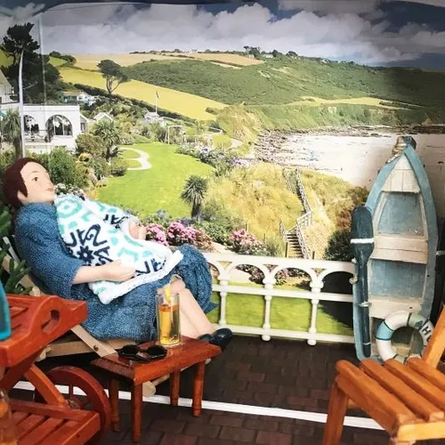 Miniature woman cradling a baby on a seaside terrace with drinks and a coastal backdrop