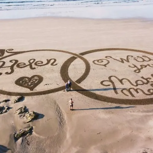 A couple stands inside a giant infinity symbol drawn in beach sand, surrounded by a heart and handwritten messages.