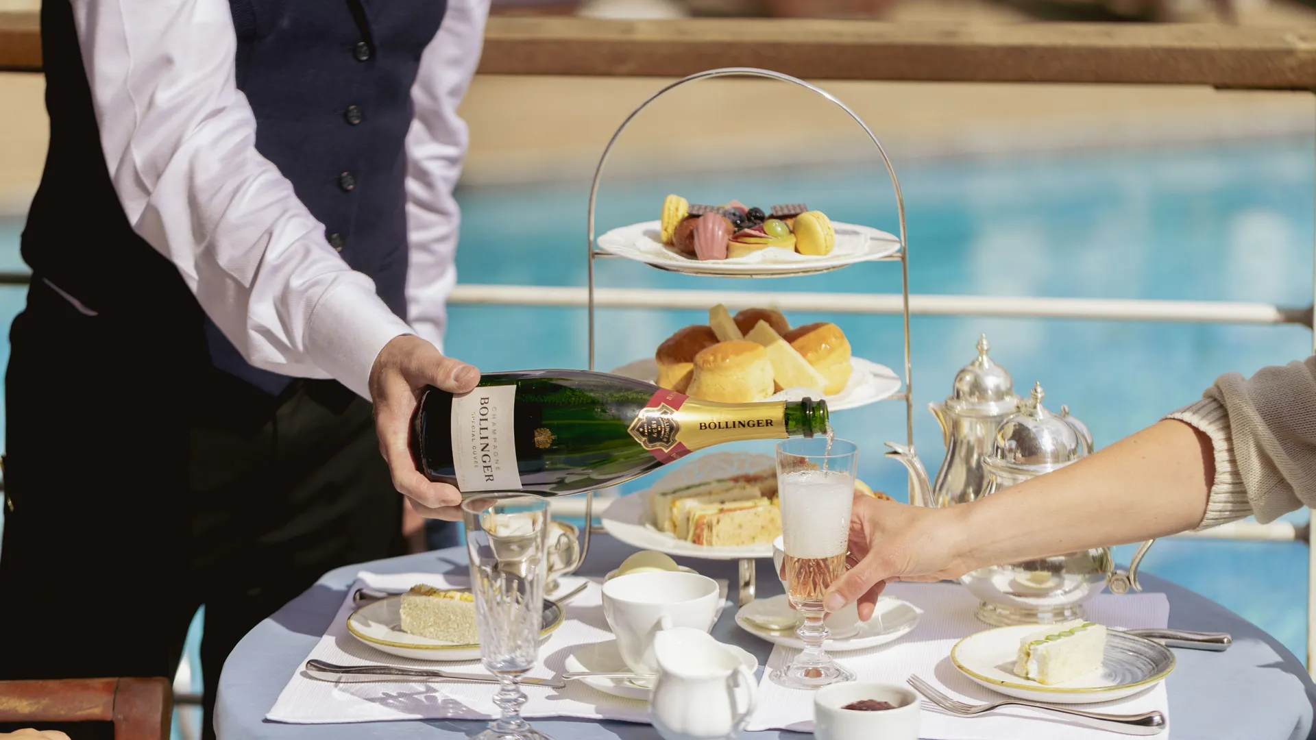Waiter pouring Bollinger champagne at a poolside afternoon tea table with pastries, sandwiches and silver teapots.