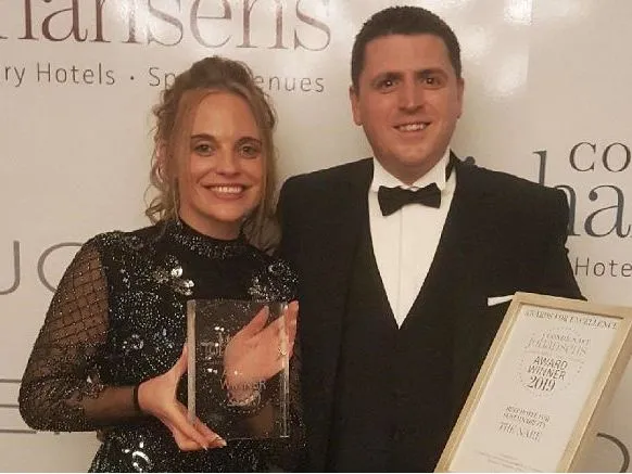 Woman in a black sequinned dress and man in a tuxedo holding an award and framed certificate