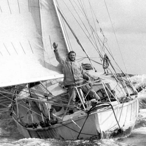 Black-and-white photograph of a sailor waving aboard a yacht sailing through choppy seas.