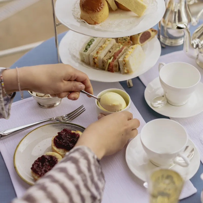 Hands preparing scones with jam and cream at an elegant afternoon tea table with sandwiches, teapots and sparkling drinks.