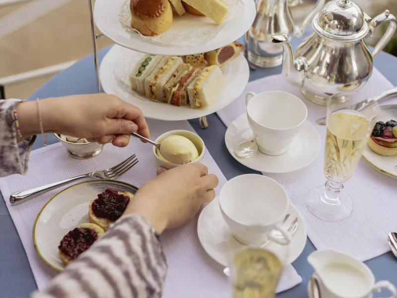Hands preparing scones with jam and cream at an elegant afternoon tea table with sandwiches, teapots and sparkling drinks.