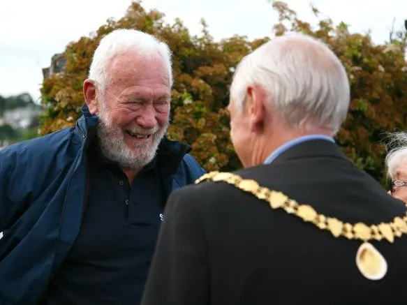 Smiling white-haired man chatting with a civic official wearing a gold ceremonial chain, with an older woman beside them outdoors.