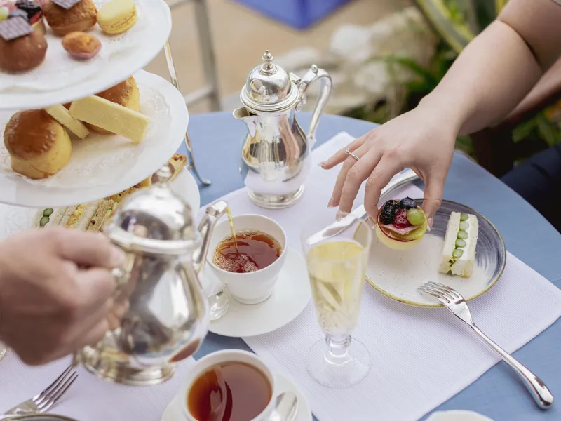 Afternoon tea with silver teapots, sandwiches, cakes and pastries on a table as a guest reaches for a fruit tart.