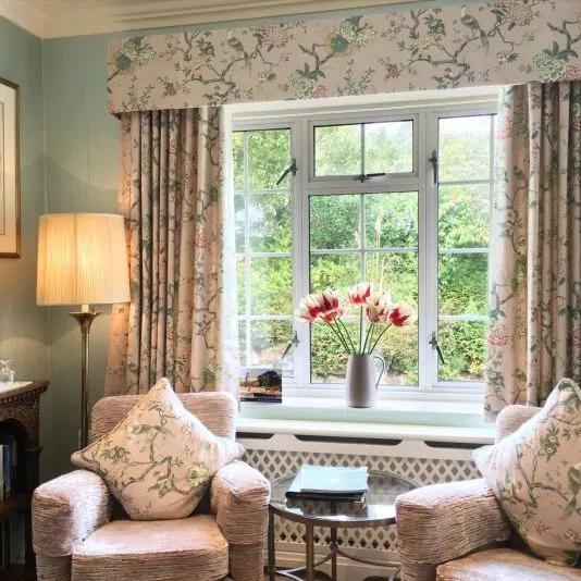 Two upholstered armchairs with floral cushions beside a window framed by patterned curtains, with flowers on the sill.