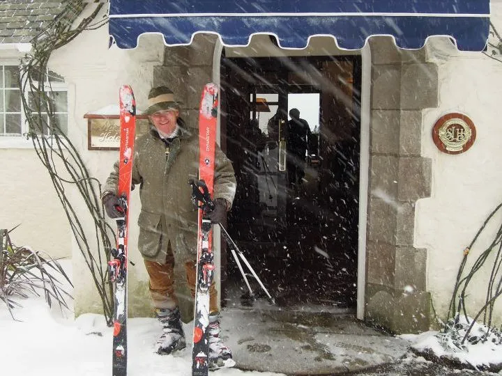 Smiling skier holding red skis outside a building during heavy snowfall