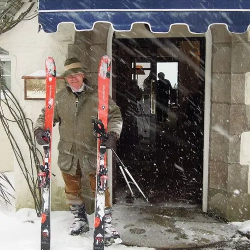 Smiling skier holding red skis outside a building during heavy snowfall