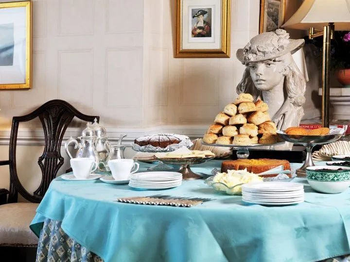 Afternoon tea spread with cakes, scones and teacups on a turquoise-covered table beside a stone bust