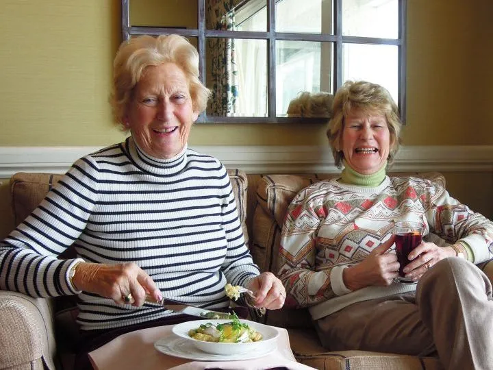 Two smiling older women sit together on a sofa, one eating from a plate and the other holding a glass of red wine.