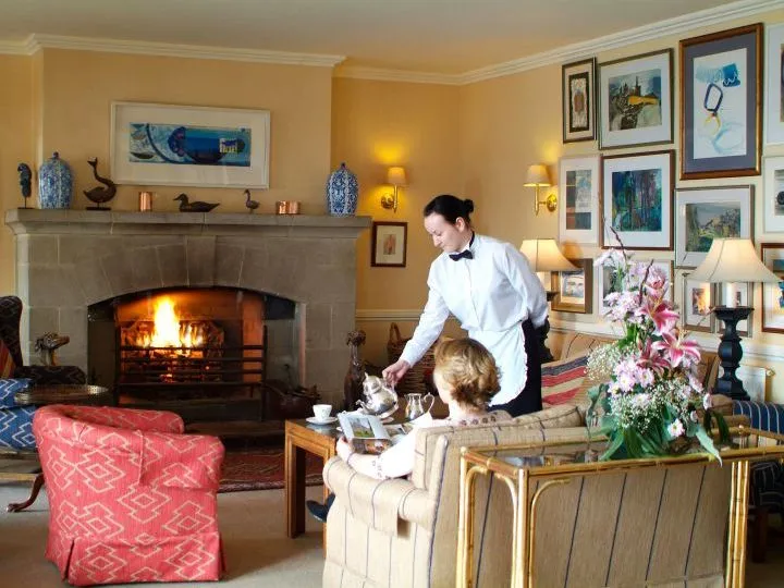 Waitress serving a seated guest beside a lit fireplace in an elegant hotel lounge