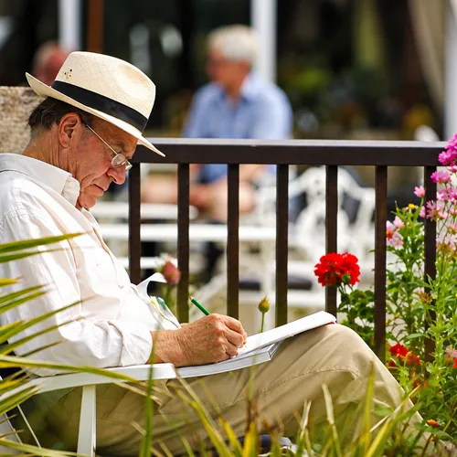 Older man in a straw hat sketching beside colourful flowers in a summer garden