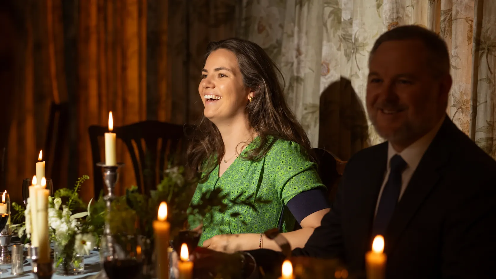 Woman in a green dress laughing beside a man in a suit at a candlelit dinner table