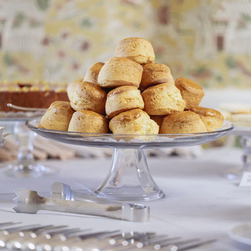 A glass cake stand piled with golden scones on a white-clothed tea table, with silver tongs and cutlery in the foreground.
