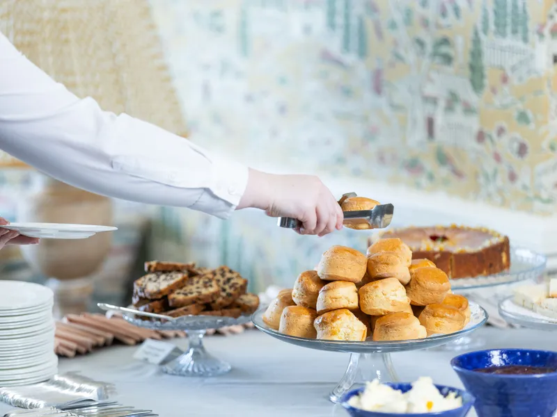 Person serving scones with tongs from a dessert table with cakes and sandwiches
