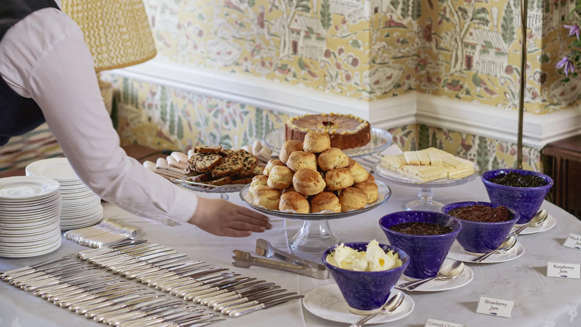 A staff member arranges a traditional cream tea table with scones, cakes, butter and jams in an elegant room with patterned yellow wallpaper.
