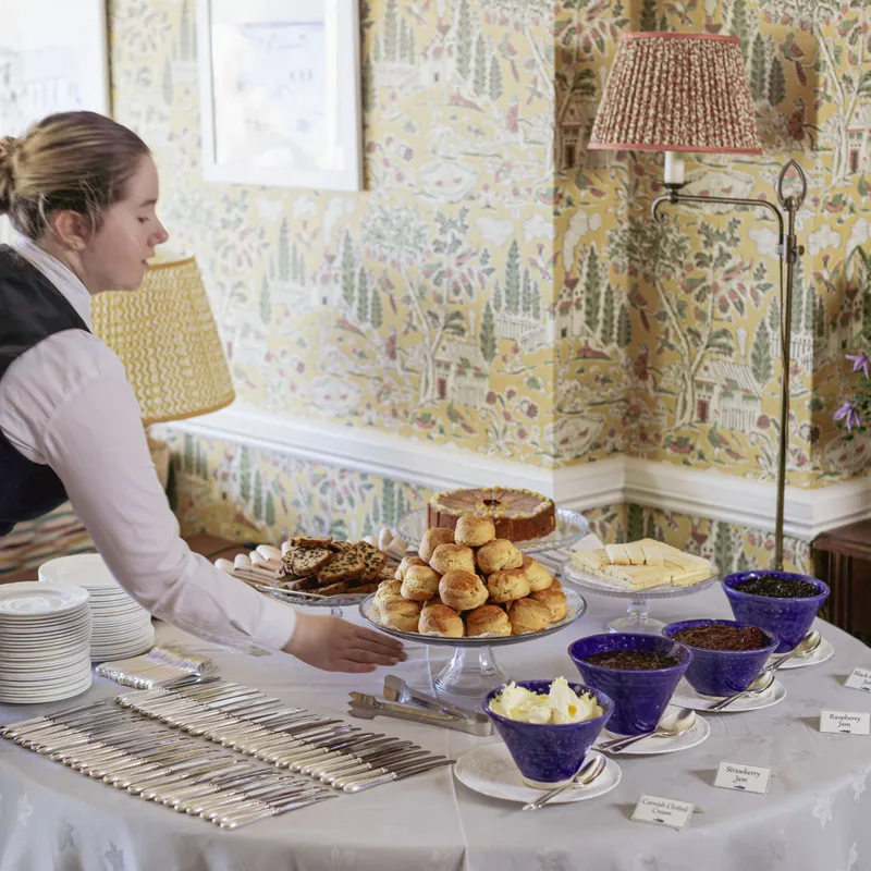 A staff member arranges a traditional cream tea table with scones, cakes, butter and jams in an elegant room with patterned yellow wallpaper.