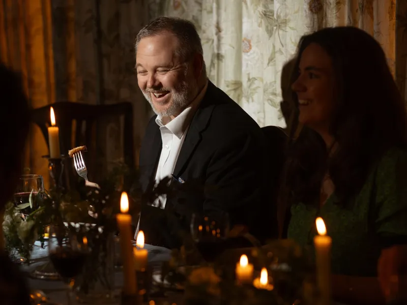 Guests enjoying a candlelit dinner at a warmly lit table, with a man in a dark jacket smiling while holding a fork.