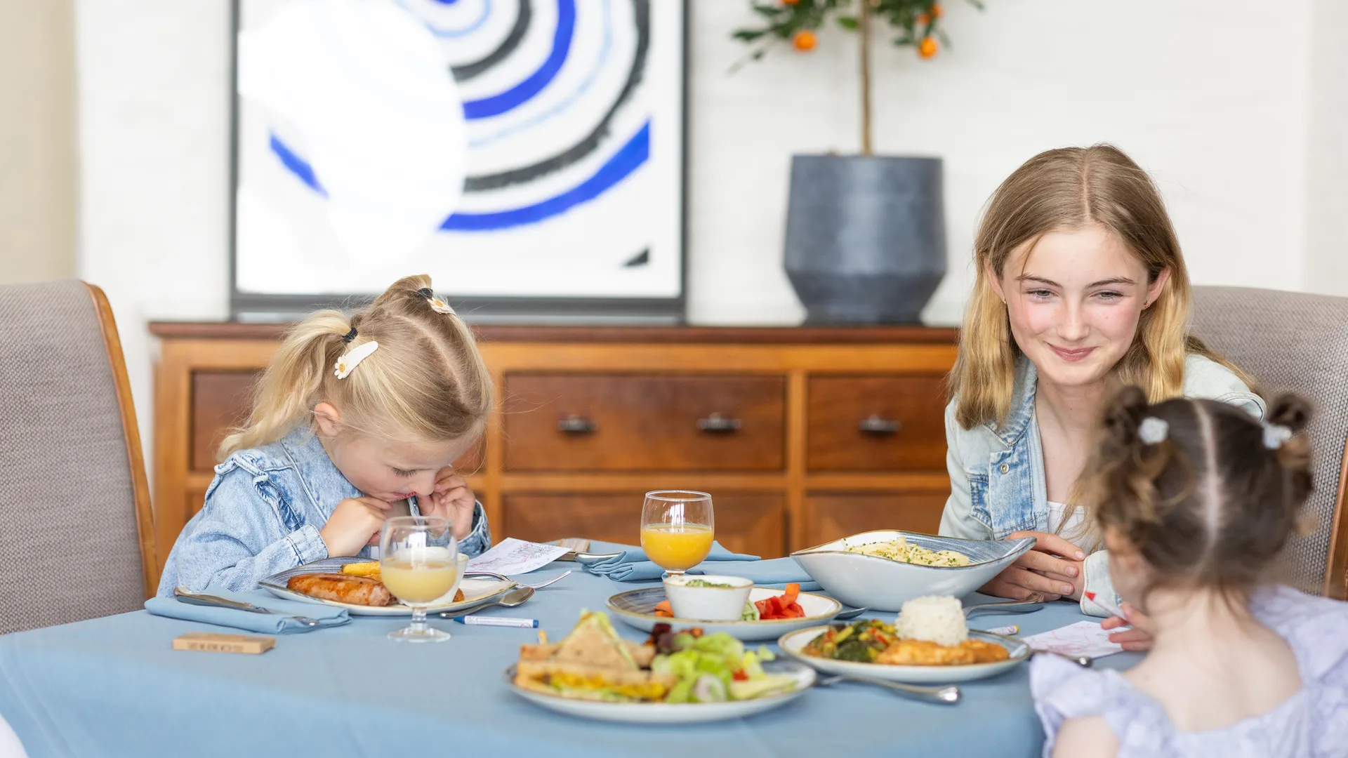 Three children sitting at a dining table with plates of food and glasses of juice in a bright, modern room.