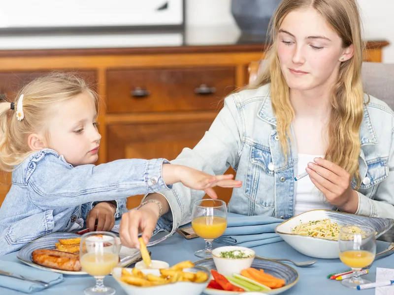 Two girls in denim jackets eating lunch at a dining table with chips, vegetables and orange juice in a bright home interior.