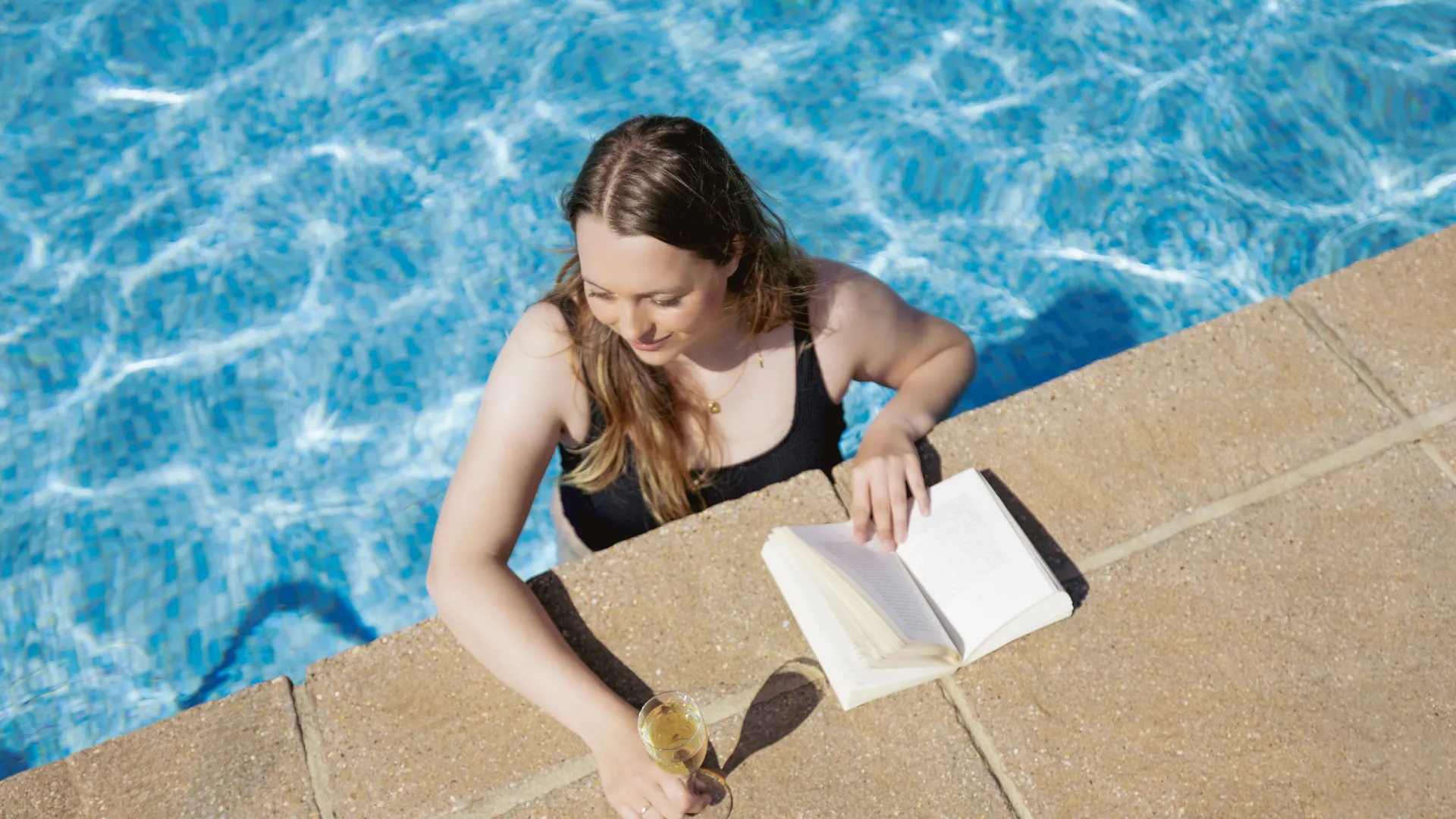 Woman in a black swimsuit relaxing in a swimming pool beside an open book and a glass of sparkling wine