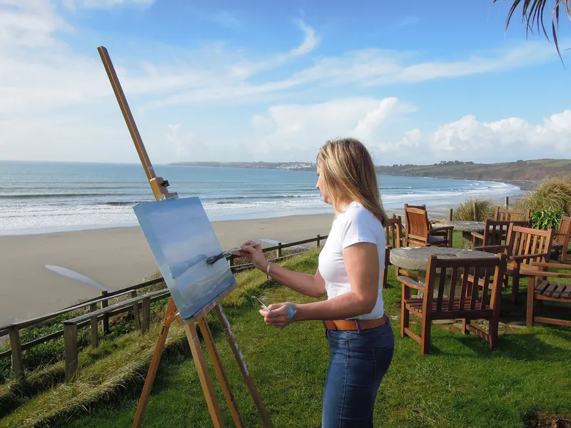 Artist painting a coastal seascape on an easel overlooking a sandy beach and calm sea