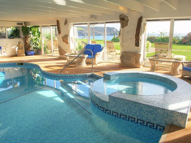 Bright indoor swimming pool with tiled hot tub, wicker seating and large windows overlooking the sea and garden