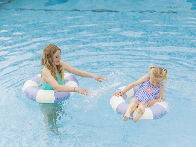 Two girls playing in a swimming pool on striped inflatable rings, splashing water and smiling.