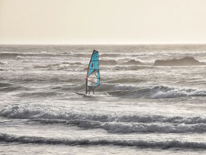 A windsurfer rides choppy waves near a sandy shore at sunset, with a bright blue sail standing out against the sea.