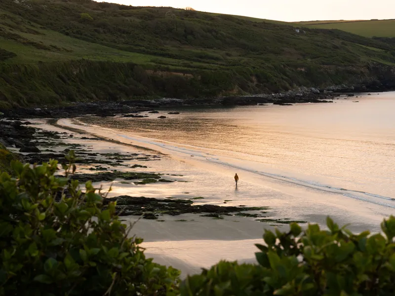 A solitary person walks along a quiet sandy beach at sunset, with calm water, rocky outcrops and green cliffs surrounding the bay.