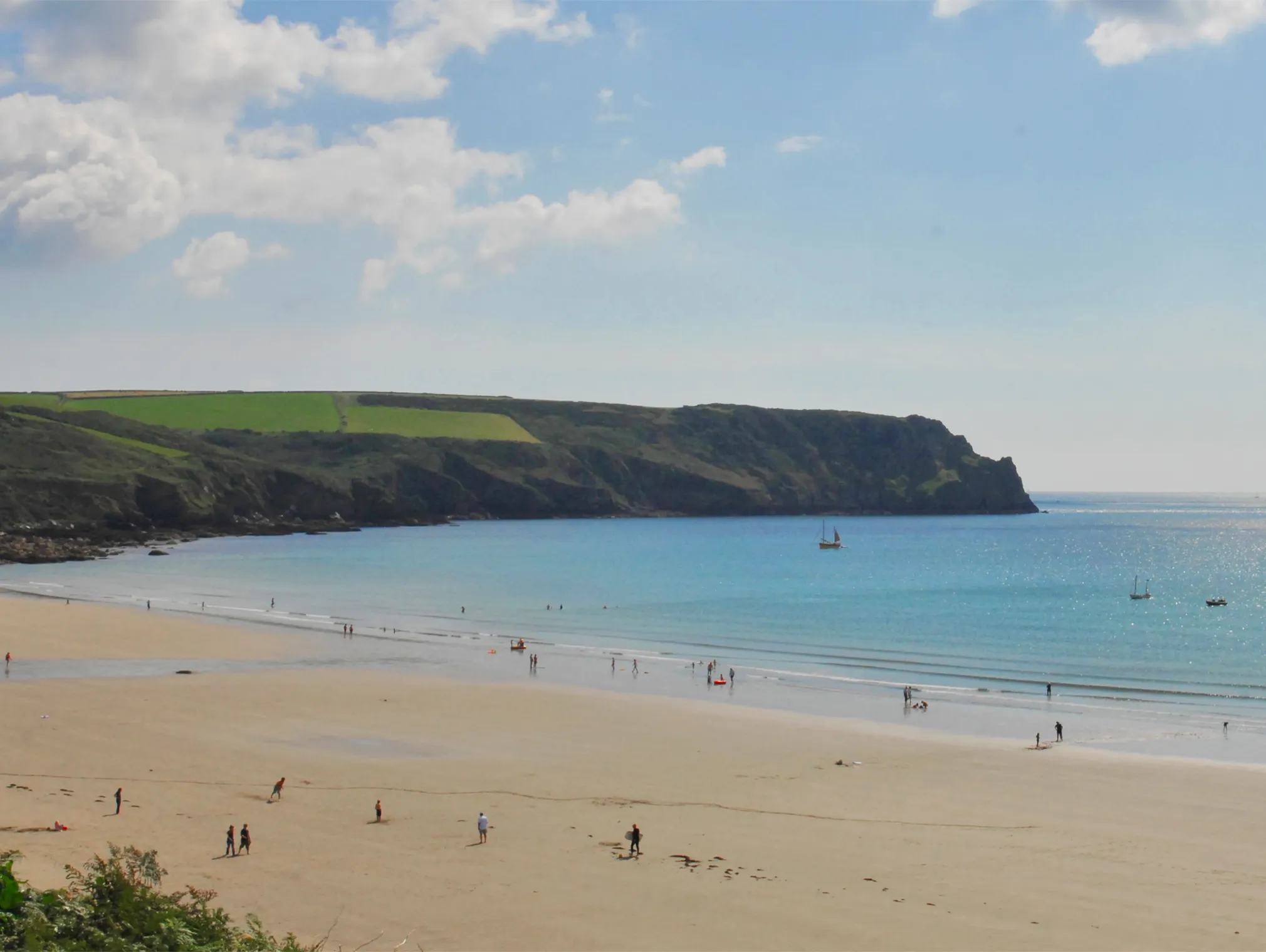 Wide view of The Nare beach with golden sand, calm turquoise sea, scattered people and boats beneath a bright blue sky.
