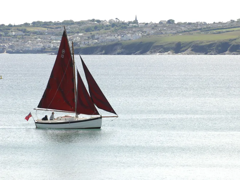 Traditional sailing boat with dark red sails on calm coastal water, with a hillside village in the background