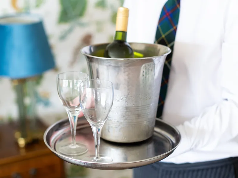 A man in a white shirt and tartan tie carries a tray with a chilled bottle of white wine in an ice bucket and two glasses.